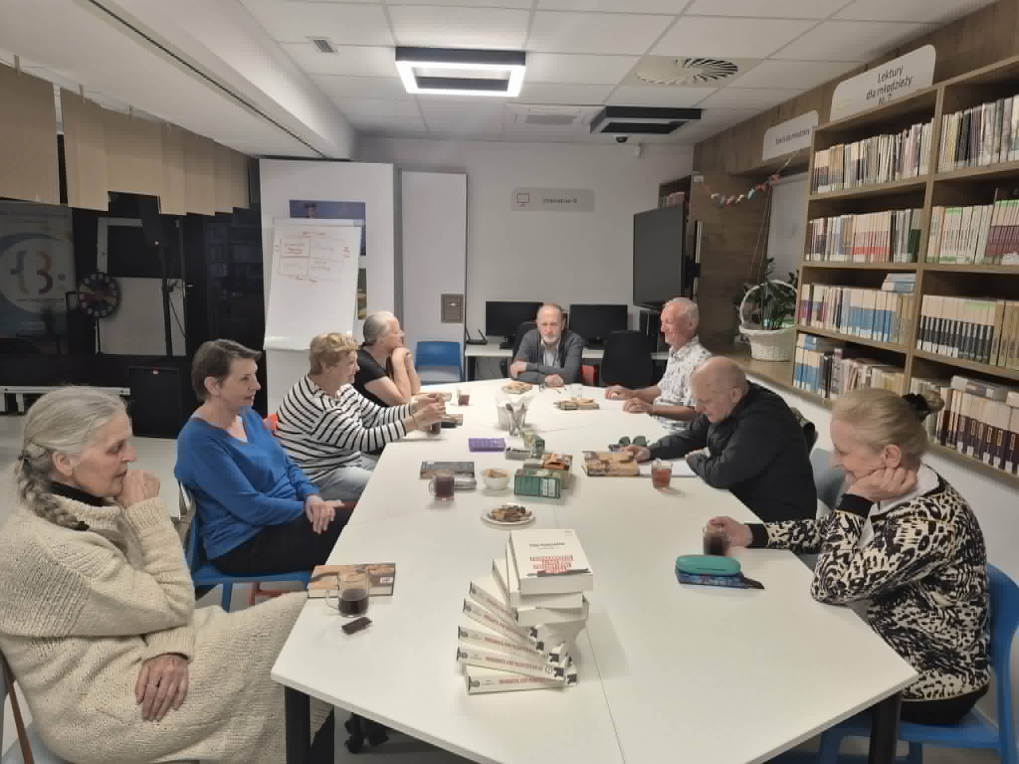 Uczestnicy spotkania klubu książki siedzą wokół podłużnego stołu w bibliotece, ułożonego w kształt litery U. Na pierwszym planie, na rogu stołu, ułożony jest wysoki stos książek. Osoby zgromadzone na spotkaniu gestykulują i rozmawiają w swobodnej atmosferze. W tle widoczne są ścienne regały z książkami oraz tablica typu flipchart