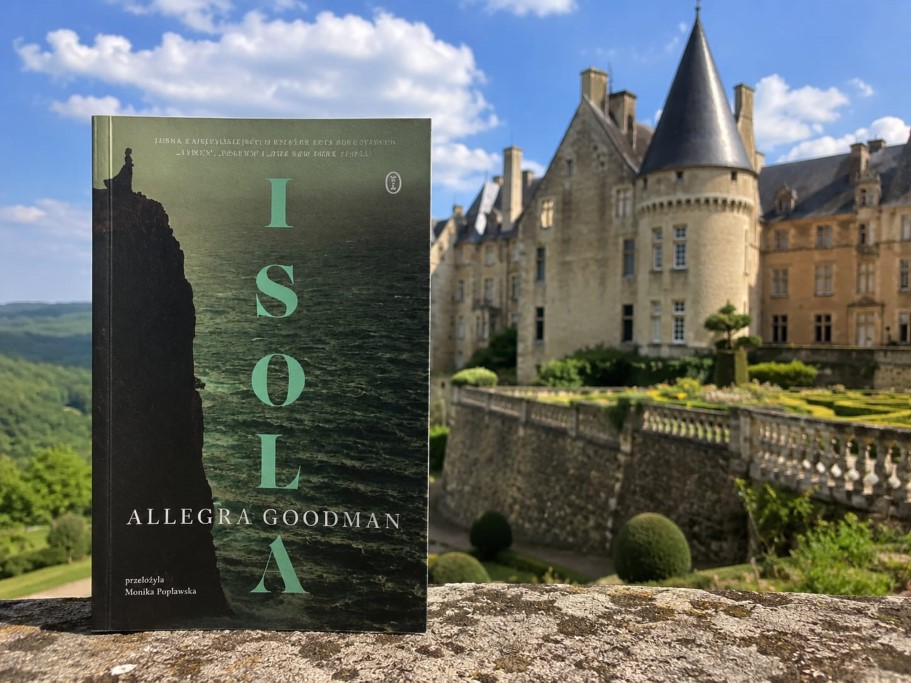 Książka „Isola” autorstwa Allegry Goodman oparta o kamienny murek. W tle rozpościera się widok na zabytkowy zamek z wieżyczkami oraz zielone ogrody i zalesione wzgórza pod błękitnym niebem. Okładka książki przedstawia ciemny klif nad wzburzonym morzem, z pionowym napisem tytułowym
