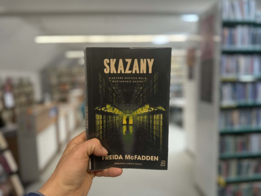 Dłoń trzyma książkę Freidy McFadden pod tytułem „Skazany” na tle rozmytego wnętrza biblioteki. Na ciemnej okładce widoczna jest grafika przedstawiająca korytarz więzienny z celami, rozświetlony żółtym światłem, na końcu którego stoi sylwetka człowieka. Pod tytułem widnieje podtytuł: „Niektóre decyzje mają dożywotnie skutki”