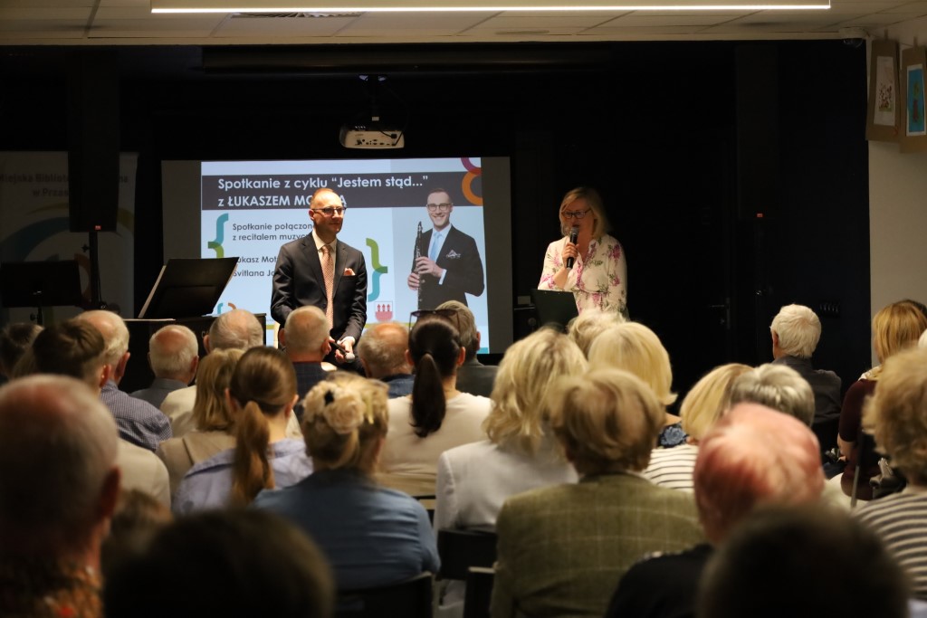 Mężczyzna w garniturze siedzi na scenie z mikrofonem w dłoni, a obok niego stoi kobieta przemawiająca do mikrofonu. Przed sceną siedzi zgromadzona publiczność. W tle ekran z plakatem wydarzenia „Jestem stąd…”