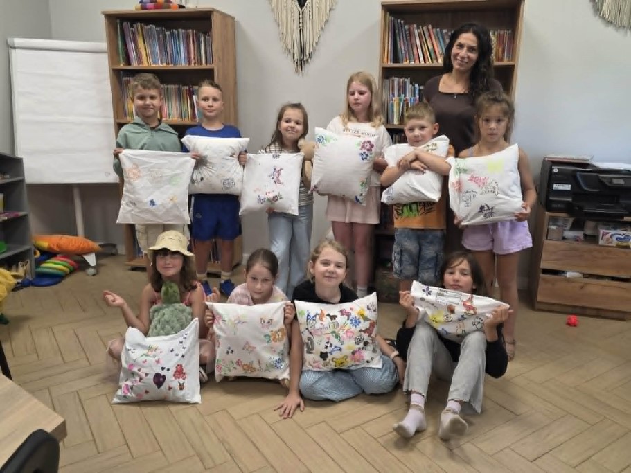 Grupa dziewięciorga uśmiechniętych dzieci oraz stojąca z tyłu ciemnowłosa instruktorka pozują w przytulnej sali bibliotecznej, prezentując białe, ozdobione kolorowymi rysunkami poduszki. W tle widoczne są regały wypełnione książkami, flipchart oraz drukarka